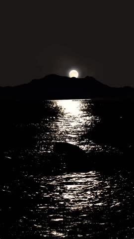 Full moon rising over dark mountain silhouettes, with bright moonlight shimmering across the rippling ocean surface. A dramatic and serene night seascape in black and white tones.

