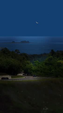 Scenic night view of a crescent moon shining over a calm ocean, with small islands visible in the distance and a curving road passing through a lush green landscape in the foreground.