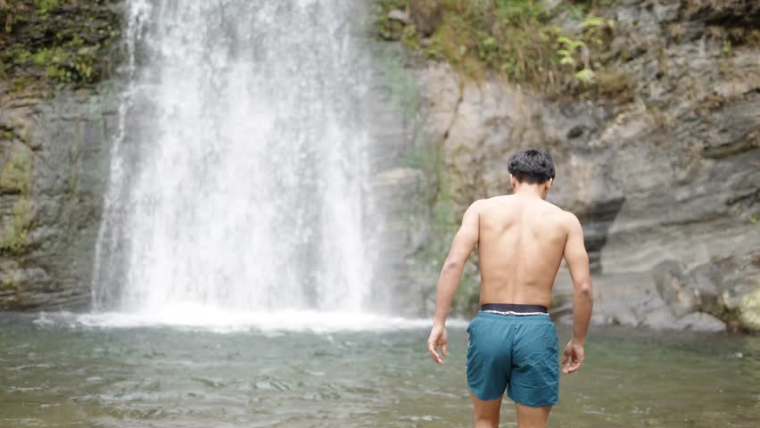 Shirtless Indian boy standing under a waterfall enjoying the water flow, raw nature moment, 4k video
