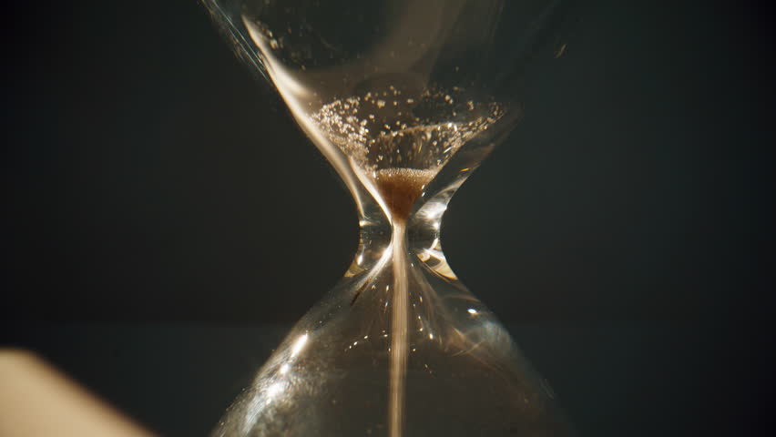 Last seconds flowing in hourglass standing at evening shadow closeup. Gold sand running inside transparent clock measuring time indoors. Elegant glass egg timer showing countdown on table surface.