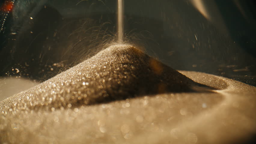 Closeup sand falling in hourglass standing at table in evening lit room. Macro view gold grains counting seconds inside elegant clock. Transparent timepiece showing time flowing. Classic decor concept