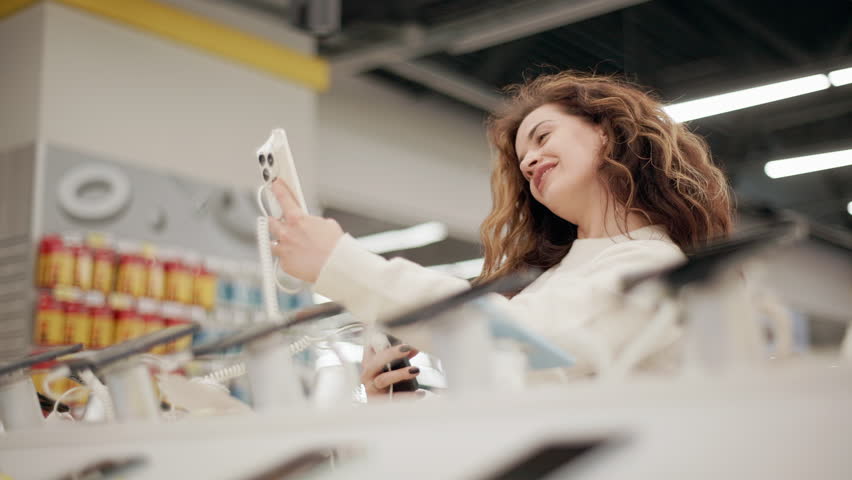 Young woman browsing multiple smartphones at electronics store, carefully comparing features, testing functionality, capturing selfies with different mobile devices in slow-motion mode
