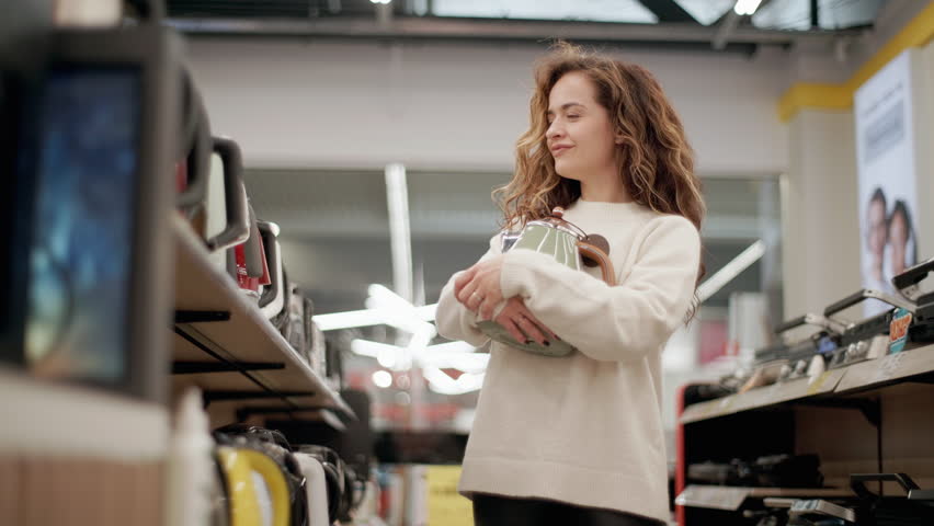 Young woman comparing kettles in an electronics store, holding a white electric kettle and a green kettle, trying to decide which one to buy, in slow motion