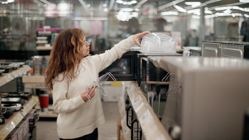Woman comparing microwave ovens and covers while browsing electronics store shelves, examining product features in slow-motion sequence highlighting consumer decision-making process