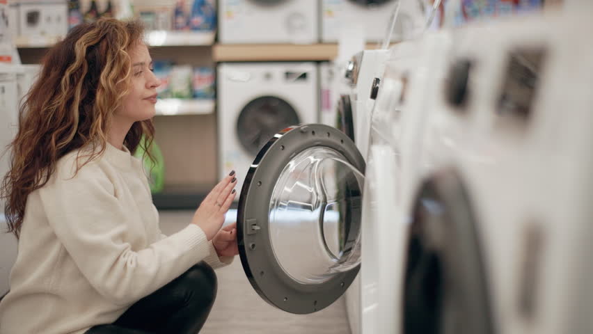 Customer carefully examining washing machines in electronics store, comparing features and specifications while browsing product display in slow motion, exploring potential home appliance purchase