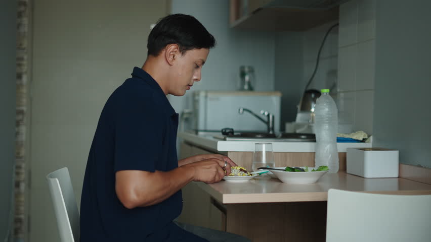 Young Asian Man Enjoying A Nutritious Meal While Sitting At A Minimalist Kitchen Table With Fresh Salad And A Glass Of Water