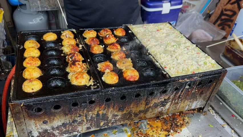 Close-up shot of Takoyaki being prepared on a hot plate. The spherical Japanese snack is almost ready, showing a delicious golden-brown color. A street food vendor expertly cooks the savory balls,