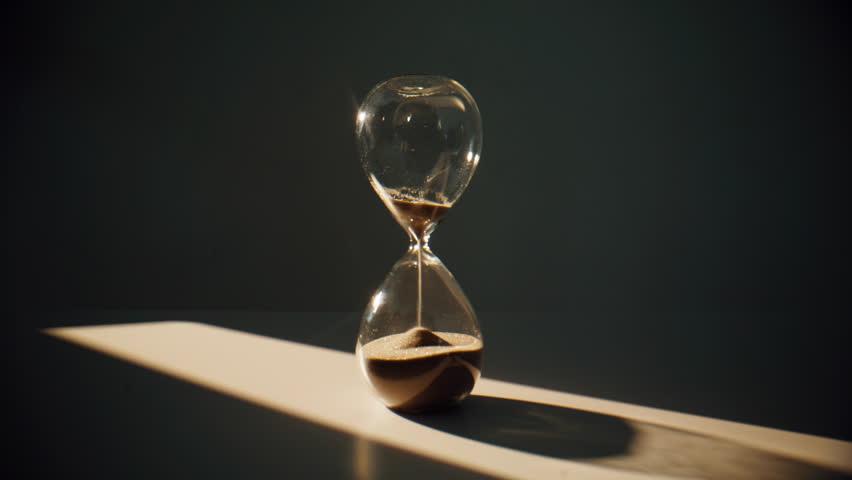 Sand flowing inside hourglass standing on surface in evening shadow closeup. Gold grains running in elegant transparent timepiece measuring minute on desk. Aesthetic clock showing time passing indoors