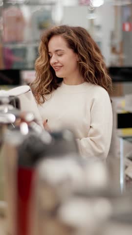 Serious young woman carefully examining electric kettle while browsing home appliance store selection, comparing product features with focused concentration