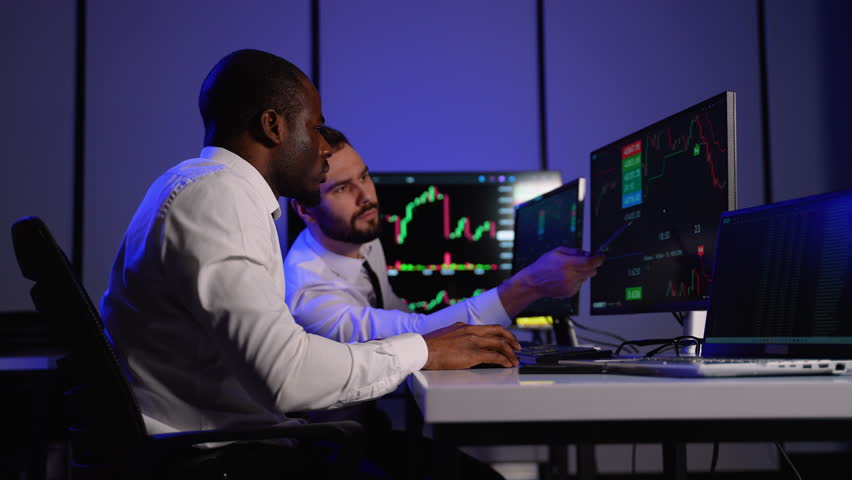Two investment fund analysts analyze stock market charts on multiple monitors in office