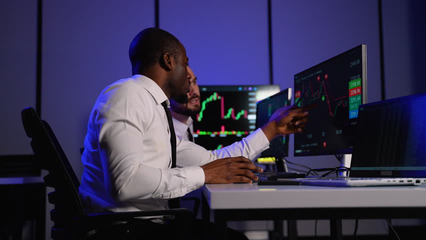 Two investment fund analysts analyze stock market charts on multiple monitors in office