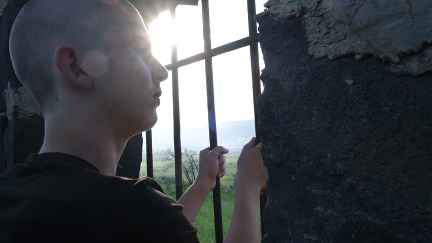 A sad bald teenage boy stands in a prison cell, holding the bars of a window and gazing at the distant mountain landscape. Concept of youth crime prevention and the consequences of poor choices.