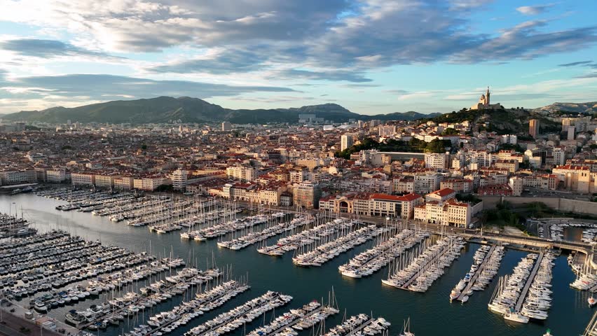 Aerial view of the Old Port of Marseille, France, with hundreds of docked yachts and boats. Historic city spreads toward the hills with the Notre-Dame de la Garde basilica visible in the background.