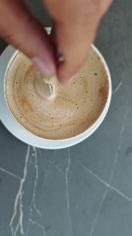 Close up of a barista stirring the creamy foam of a cappuccino with a teaspoon, creating a delicate swirl