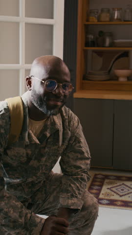Medium vertical shot of happy African American marine dad in khaki military uniform, with backpack kneeling in doorway after returning home from mission and hugging excited teenage son