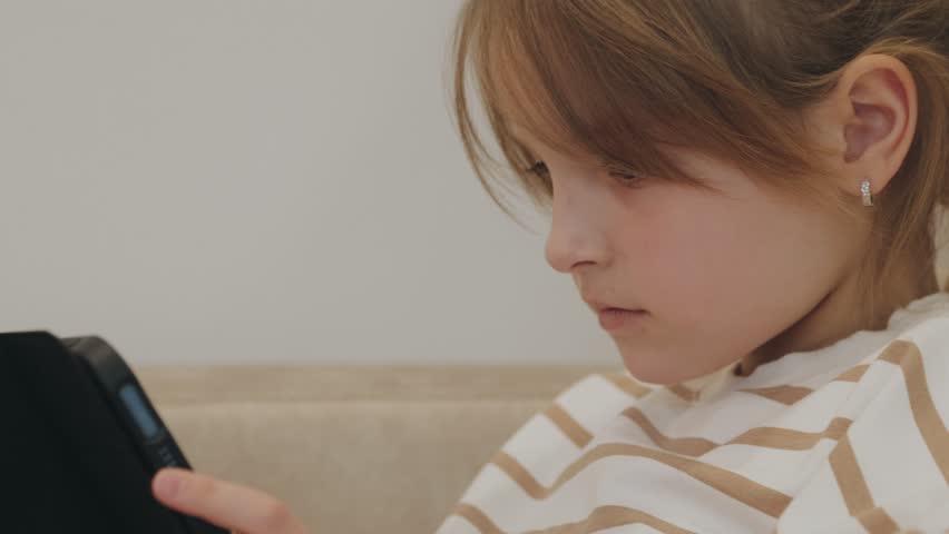 Focused young girl using a tablet, engrossed in educational content, comfortably seated at home, possibly reading or completing online lessons