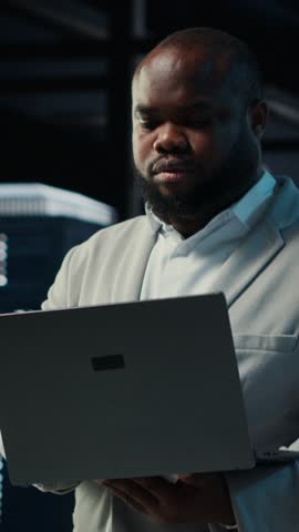 Vertical video Data center admin coding, writing script on laptop, identifying bottlenecks. Server farm IT expert typing on notebook keyboard, troubleshooting bugs, implementing optimizations, camera