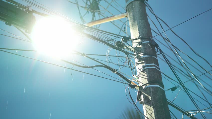 Close-up of an electric pole and tangled wires during rain, backlit by intense sunlight.