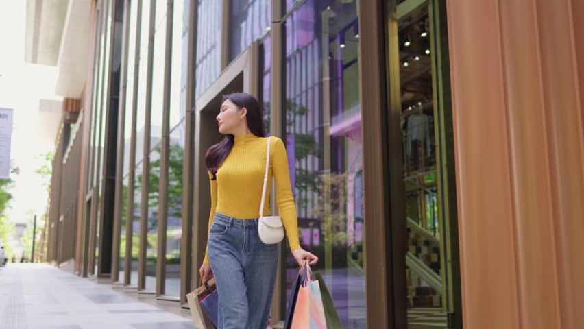 Asian young beautiful woman shopping goods outdoor in department store. Attractive female holding shopping bags then walking alone with happiness, enjoy purchasing in shopping mall marketplace center.