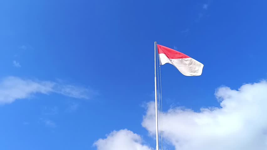 The Indonesian national flag flutters against a cloudy blue sky background from a low angle camera. The red and white flag. The Unitary State of the Republic of Indonesia.