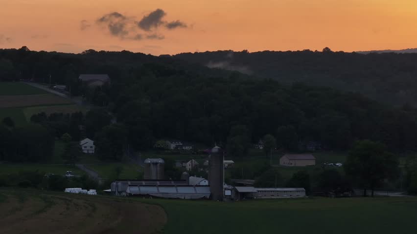 Aerial wide shot of american farm with silo storage during golden sunrise. Yellow lighting sky behind mountain and hills of America. Dusk scene in rural district of city.