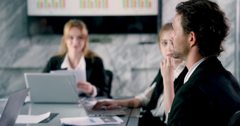 Caucasian male adult employee listening carefully during office meeting with colleagues exchanging ideas using laptops discussing teamwork strategy and presenting data on large wall chart behind team