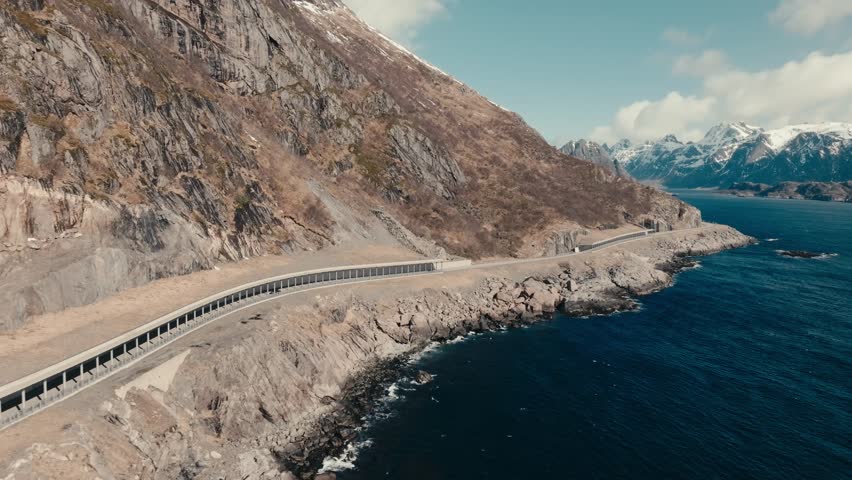 Lofoten Islands, Nordland, Norway - Cars Glide Along a Dramatic Coastal Road, Flanked by Rugged Cliffs and the Deep Blue Sea - Aerial Drone Shot