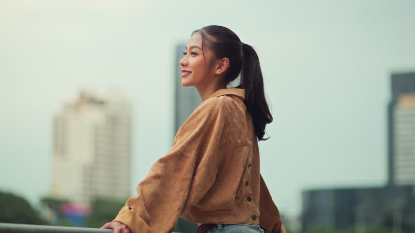 Cheerful asian woman standing on bridge in city park enjoying fresh air and nature – happy female smiling and breathing deeply in urban green space, mindful wellness lifestyle and outdoor relaxation
