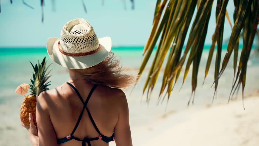 Woman in bikini and straw hat holding a pineapple cocktail on a tropical beach. Sunny summer day, turquoise ocean, palm leaves, and vacation vibes perfect moment of exotic relaxation and travel