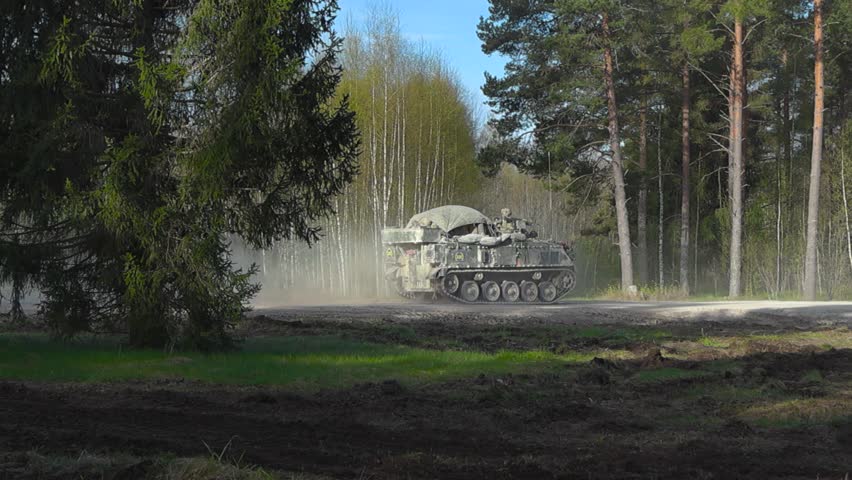British military Bulldog army armoured personnel carrier driving on a gravel dry road in a sunny forest with a massive dust cloud behind it due to high speeds. Armed soldiers are on top of the tank.
