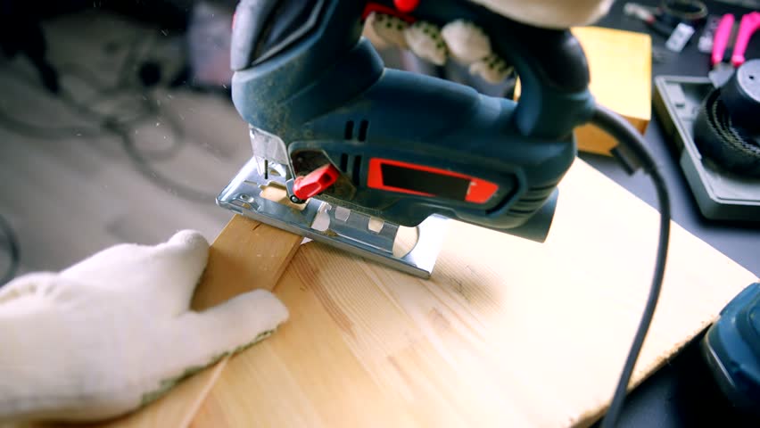 worker using electric tool for cutting wood. gloved hand using an electric jigsaw to cut wooden board in a workshop. DIY woodworking project in progress, with tools and equipment. cutting wood diy 
