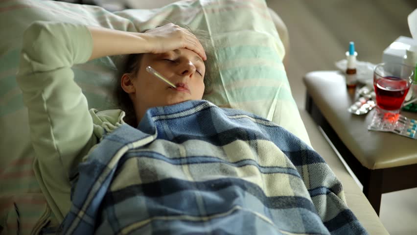Sick woman lying in bed under a plaid blanket with a thermometer in her mouth, hand on forehead. Pills, nasal spray, and hot tea on table nearby. Concept of flu, fever, or seasonal illness recovery