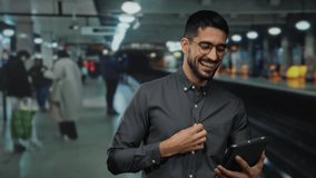 Young man using a tablet in a busy indoor train station, smiling, wearing glasses, with people in the background, illustrating modern travel and communication. - Powered by Shutterstock - Get 15% off with code: PIKWIZARD15