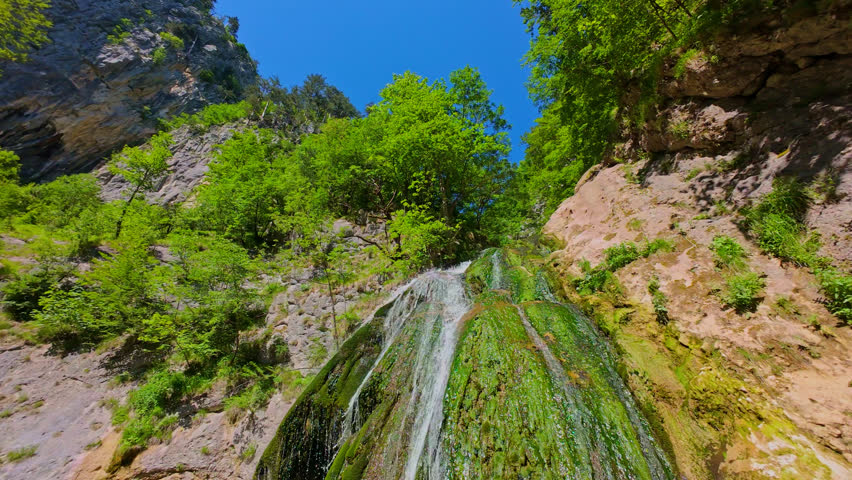 Aerial view of a scenic waterfall cascading down a rocky mountain cliff surrounded by lush green trees and forest, captured in smooth FPV drone slow motion on a sunny summer day, perfect for nature
