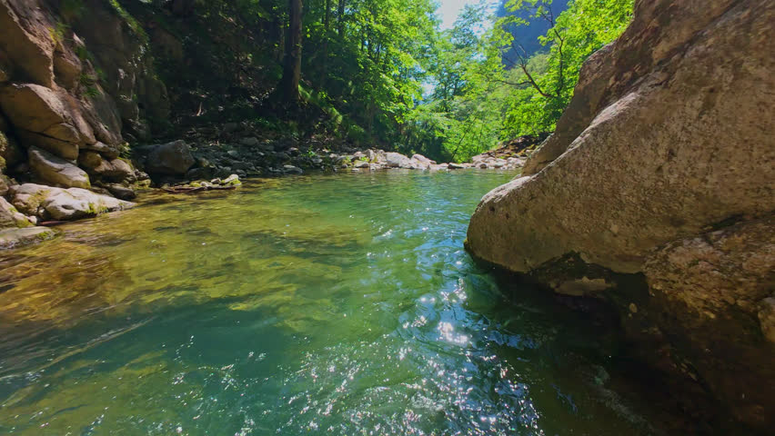 Aerial view of a scenic waterfall cascading down a rocky mountain cliff surrounded by lush green trees and forest, captured in smooth FPV drone slow motion on a sunny summer day, perfect for nature