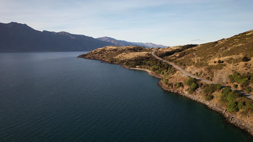 Coastal Road Along Golden Hills On Shore Of Lake Hawea In Otago Region, New Zealand. Mountain Range In Background. aerial pullback shot