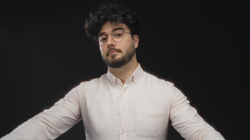 Young man with beard and glasses making air quotes gesture against a black background, showcasing expression and gesture in a studio setting.