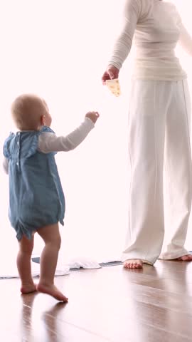 Mother encouraging baby to take first steps at home in bright room. Candid family and toddler development, early childhood milestone, parenting. Mother and child learning to walk indoors