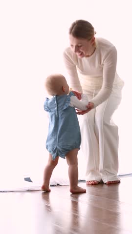 Mother encouraging baby to take first steps at home in bright room. Candid family and toddler development, early childhood milestone, parenting. Mother and child learning to walk indoors