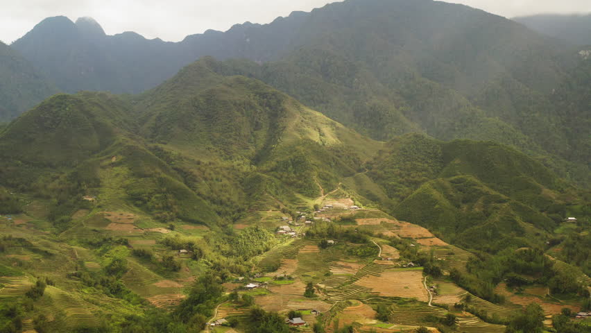 Drone orbits in Sa Pa - Ta Van above mountain landscape with terraced farmland and small homes below, as sunbeams filter through clouds over highlands near the cloud creaks and funicular train route.