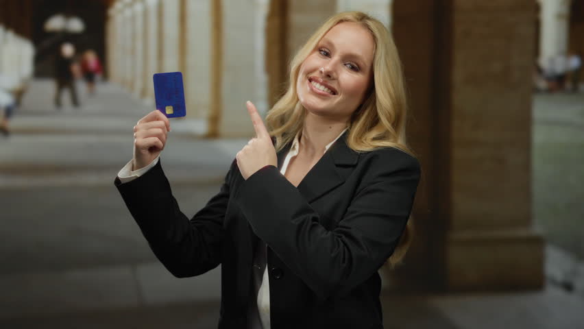 Woman showcasing credit card in university campus corridor, exuding confidence and style, surrounded by historic architecture, embodying modern education and finance.