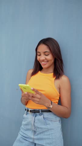 Vertical video of Young Latin woman using a smartphone,texting and talking leaning on a blue wall
