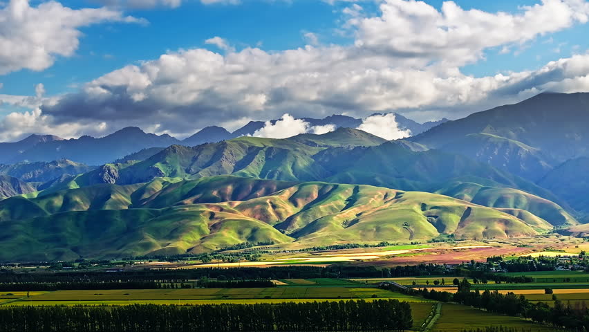 Aerial shot of rolling green hills and agricultural fields with majestic mountain range natural landscape in Xinjiang, China.