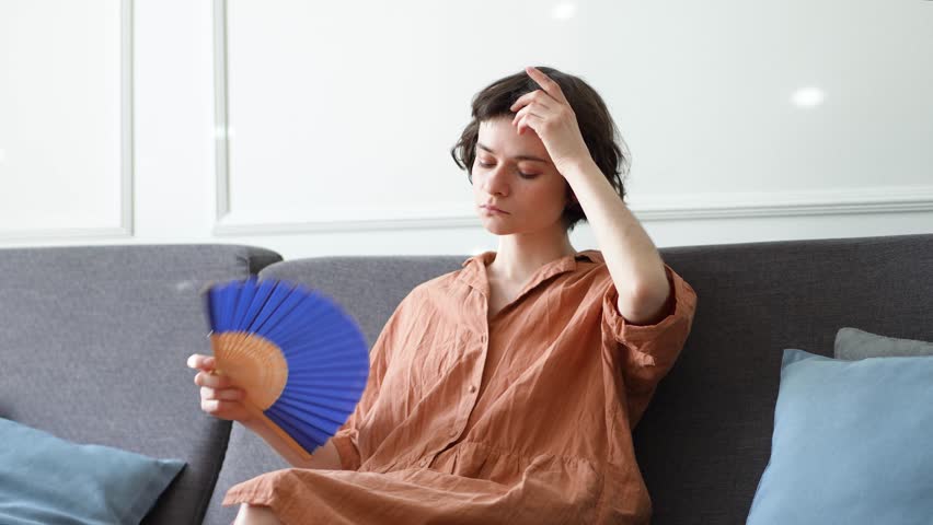 Exhausted sweaty woman fanning herself on sofa suffering from heat at home without AC. Female enduring high temperature discomfort, stuffy conditions causing dizziness and headache, summer distress