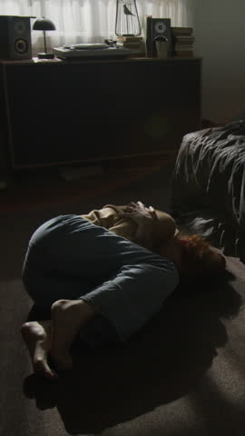 Vertical shot of vulnerable unrecognizable woman lying curled on floor near bed in dimly lit unkempt room