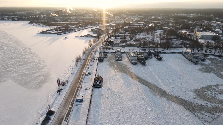 An aerial view of a snow and ice covered port with several ships docked, set against a dramatic winter sunset over the frozen bay