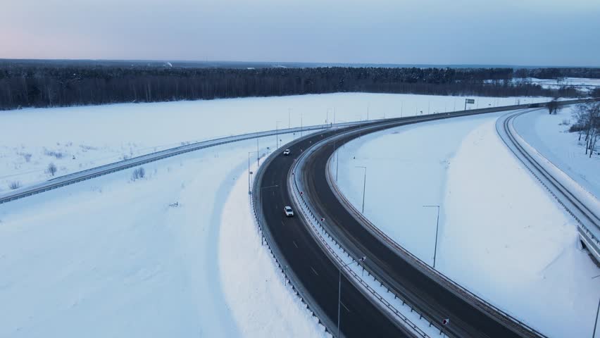 An aerial view of a highway and a bridge traversing a snow covered winter landscape under a bright sunset sky