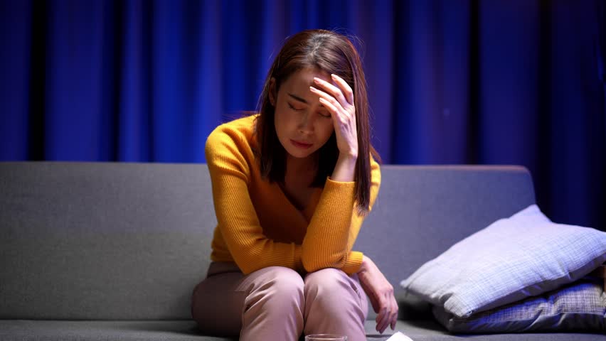 Video 4k of Asian woman is resting on the sofa looking unwell with a headache and fever. She appears stressed tired and exhausted from cold flu or illness at home.