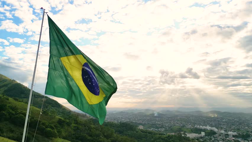 Large Brazilian Flag Waving in the Sky With Nova Iguacu City in the Horizon and the Sun Shining Through Clouds