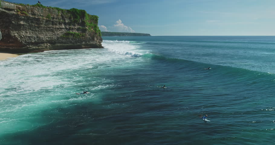 Turquoise ocean waves rolling onto Pantai Dreamland Beach in Bali, with surfers eagerly waiting to ride the waves against a backdrop of cliffs and lush vegetation